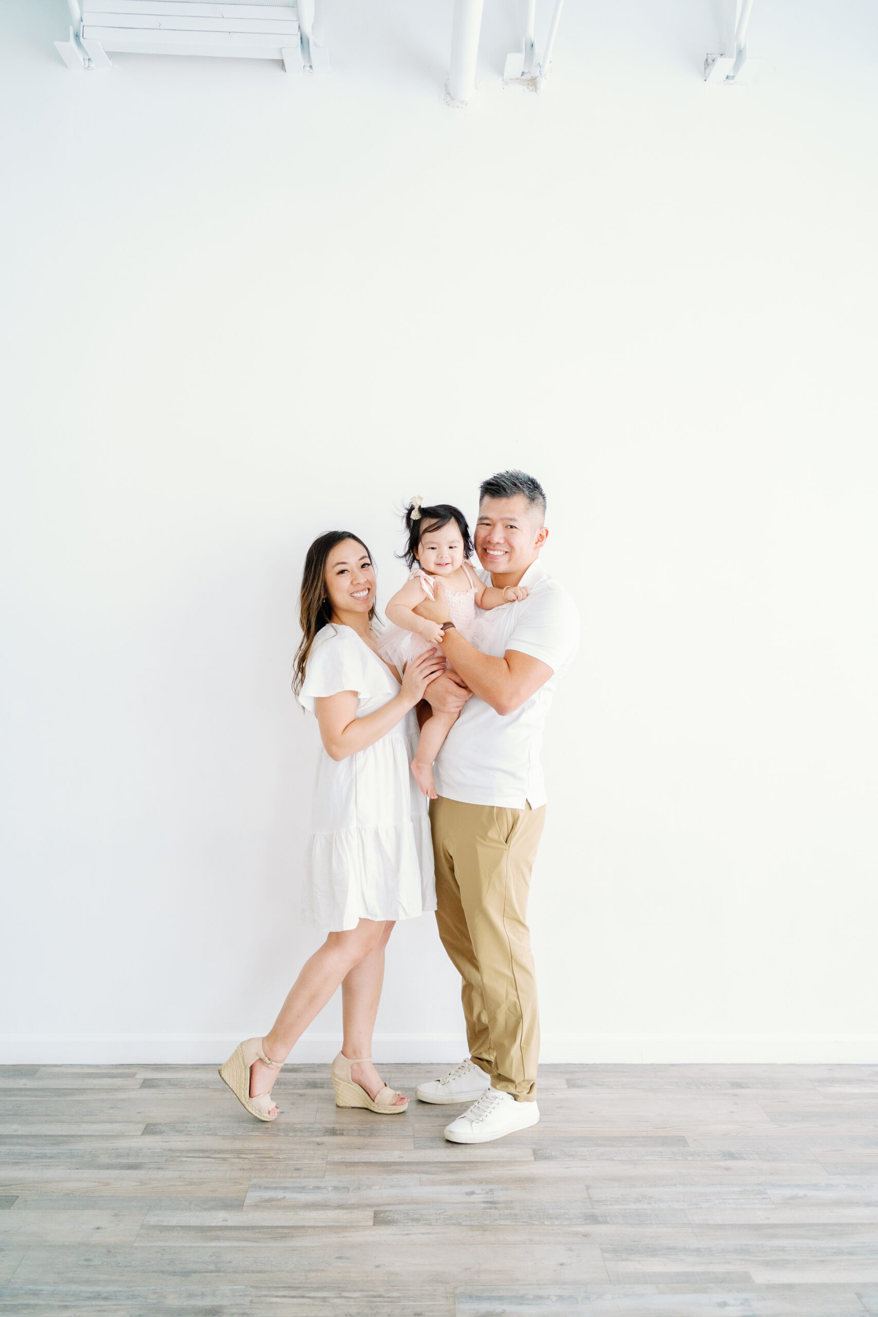 Family portrait with parents holding their baby, standing in front of a simple neutral backdrop.